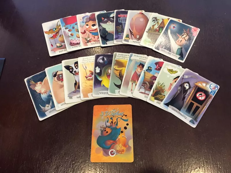 3 Wishes 游戏所有卡牌的平铺展示图，上方并排陈列着多种愿望卡，下方正中是印有神灯精灵的亮黄色游戏封面卡，整体插画风格生动且富有童趣。